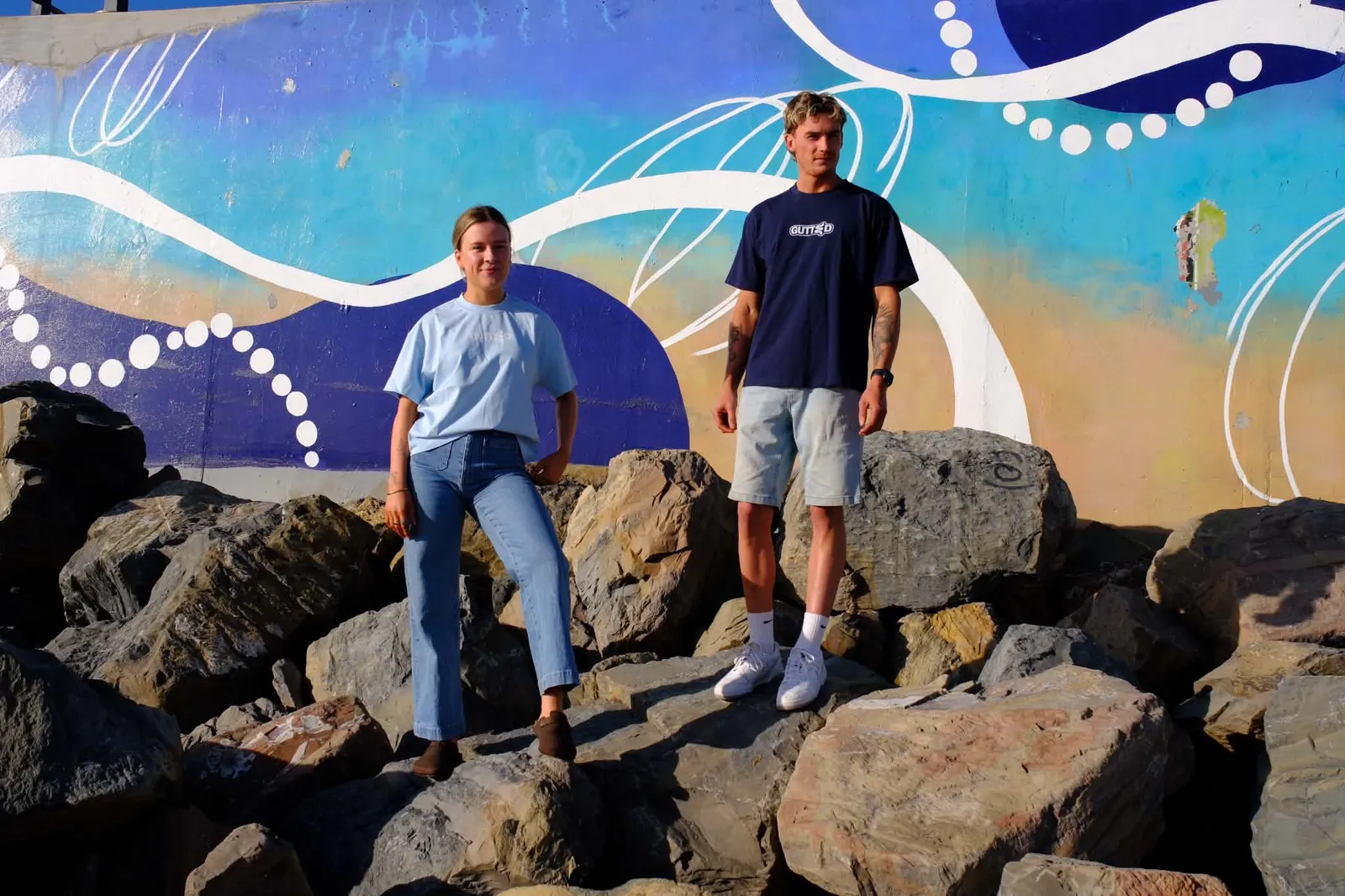 Two people wearing Gutted tees in front of a colourful mural